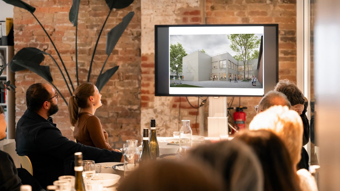 Team von Sehw Architektur beim Aperitivo, sitzend am Tisch und gemeinsam auf die Präsentationsleinwand blickend.