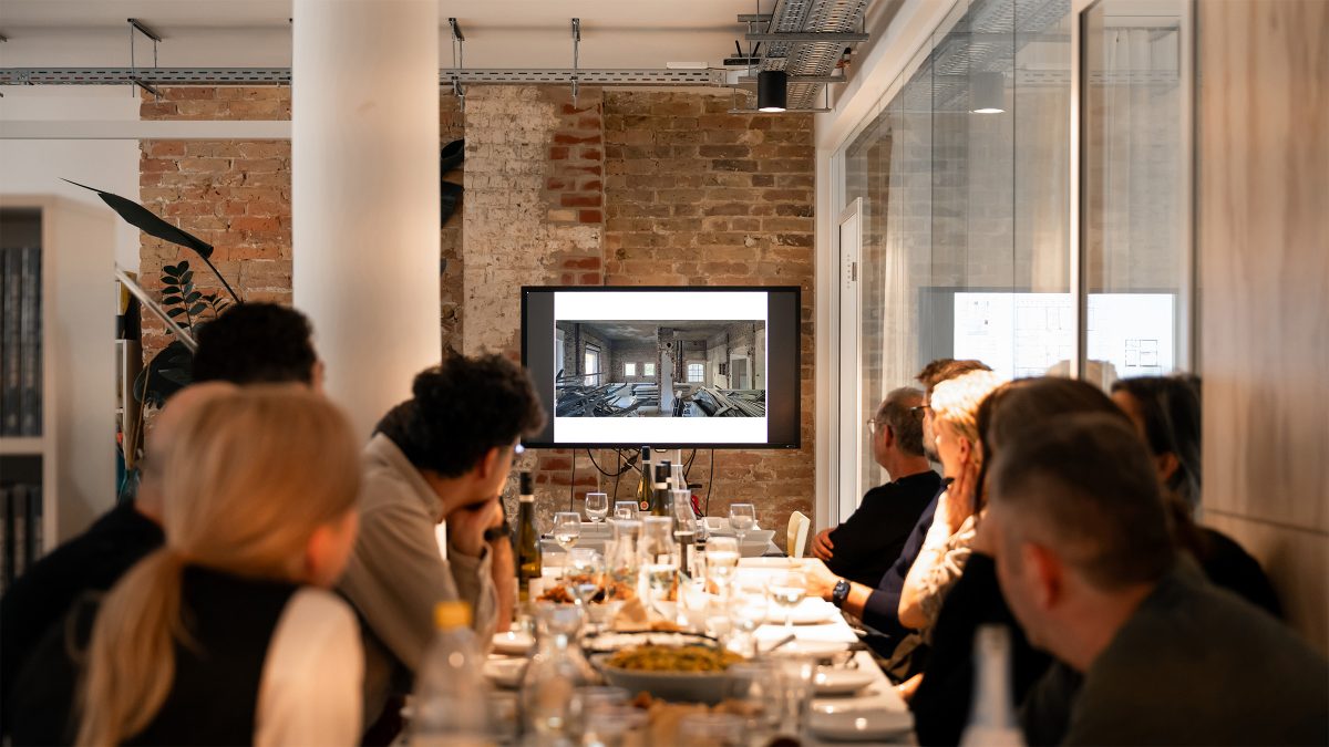 Team von Sehw Architektur beim Aperitivo, sitzend am Tisch und gemeinsam auf die Präsentationsleinwand blickend.