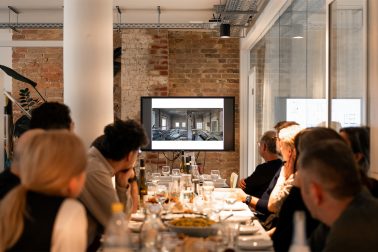 Team von Sehw Architektur beim Aperitivo, sitzend am Tisch und gemeinsam auf die Präsentationsleinwand blickend.