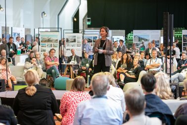 Oya Sönmez steht im Zentrum des Bildes und hält eine Rede während der Heinze ArchitekTOUR PROJECTS‘25, vor dem Rochadegebäude in Essen, einem dreigeschossigen, modularen Pavillon, der als temporärer Lernort für Schulen dient.